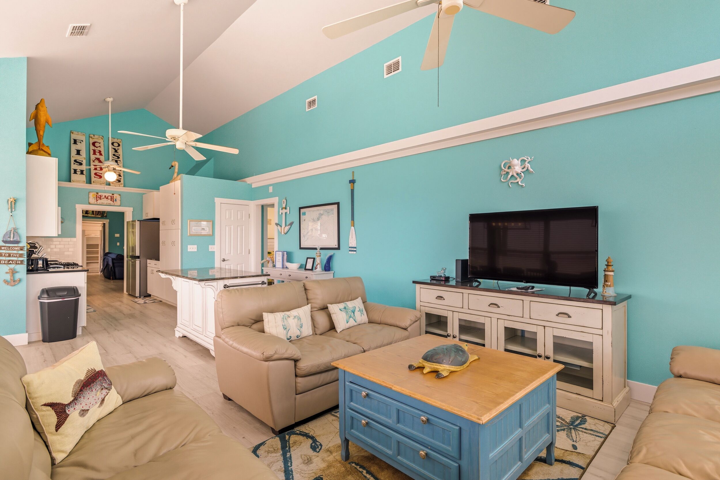 Una sala de estar con pared verde azulada, sofá color beige, aparador azul y televisor.
