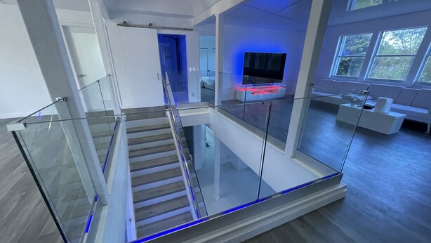 Un interior moderno con escalera de barandilla de vidrio, televisor de pantalla plana y zona de estar con vistas al exterior.