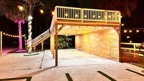 Una terraza de madera con una escalera que lleva a una zona cubierta, rodeada de luces colgantes y palmeras.