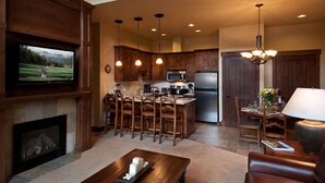 Amplia sala de estar con chimenea, muebles de madera, televisor y una zona de cocina con alacenas de madera y electrodomésticos modernos.