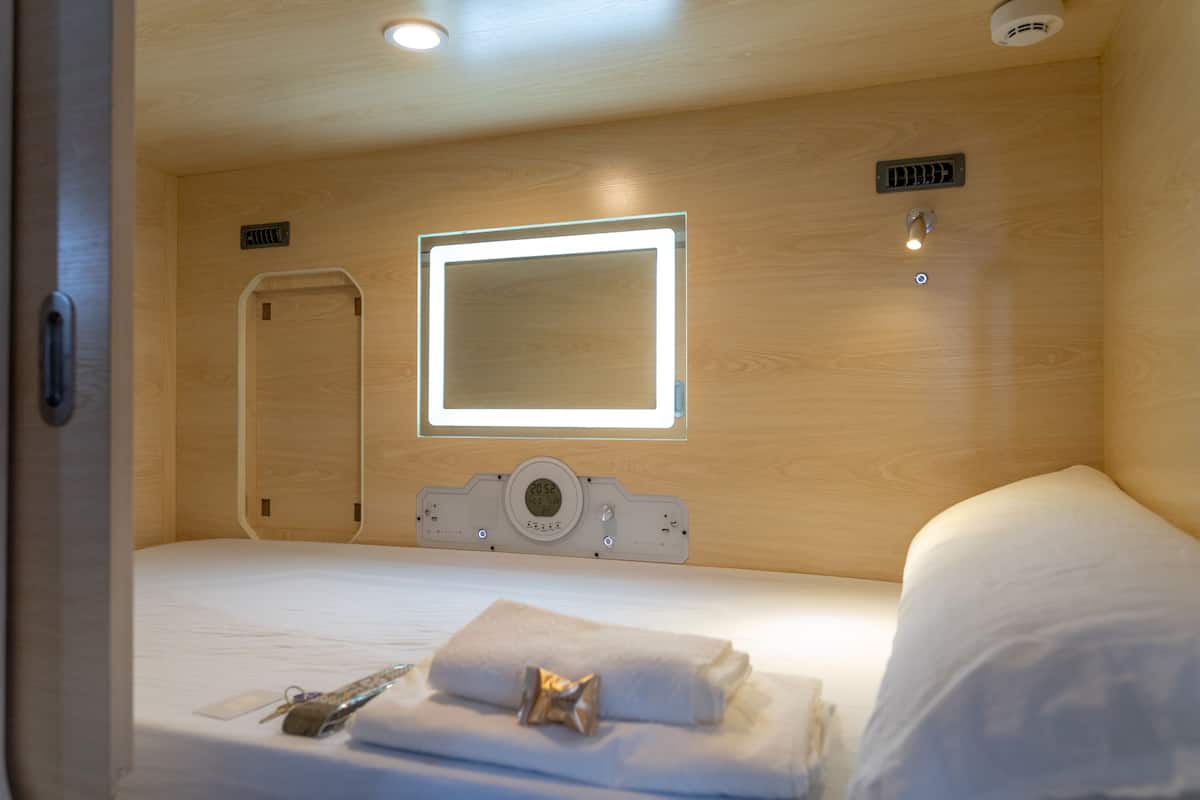 Quarto de hotel com cama, mesa de cabeceira, espelho e relógio fixado na parede.
