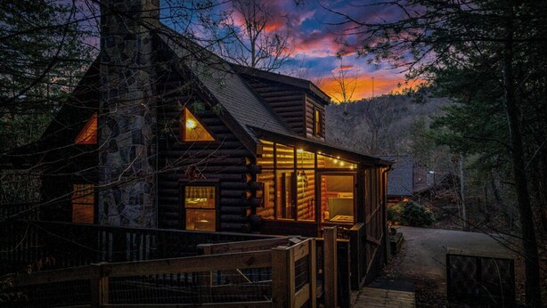 A log cabin with a stone chimney, a porch, and a view of the surrounding forest. - Timeless Tranquility-Screened Hot Tub-5 Min to Parkway-Pet Paradise-Dog Run! (Pigeon Forge)