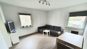 A modern living room with a grey sofa, a small white table, a TV on a cabinet, and two windows with plants. - Apartment Pottmensch near Centro with parking lot (Oberhausen)