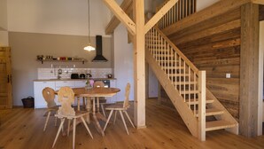 Een houten trap die leidt naar een hoger gelegen gedeelte, een keuken met witte kasten en houten stoelen, en een eettafel met stoelen.