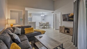 A modern living room with a sectional sofa, a coffee table, and a TV. There is a dining area in the background with a chandelier. - Newly renovated, free WIFI/PARKING, 10 min to the beach! (Clearwater)