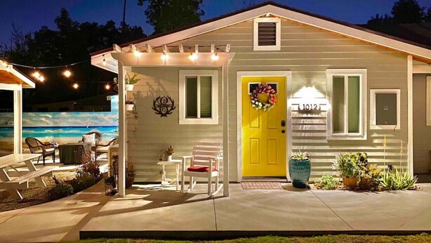 Una casa con puerta amarilla, un porche cubierto con luces colgantes, piscina y mobiliario de exterior.