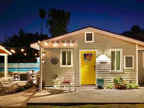 Une maison avec une porte jaune, une véranda couverte décorée de guirlandes lumineuses, une piscine et un espace salon extérieur.