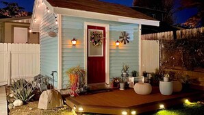 Una casita azul claro con una puerta roja, un porche con plantas en macetas y una valla de piquetes blancos.