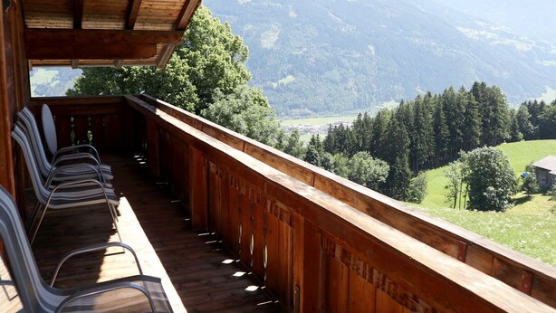 Un balcone in legno con sedie in metallo, affaccia su un paesaggio montuoso con alberi e un edificio in lontananza.
