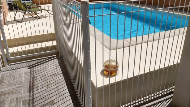 A swimming pool surrounded by a white metal fence, with a tiled deck and a chair visible in the background.