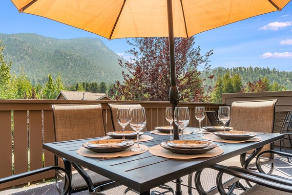 Un espace extérieur aménagé pour les repas, avec une table dressée pour six personnes, entourée de chaises, protégée par un grand parasol, et offrant en arrière-plan une vue panoramique sur les montagnes.
