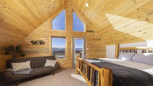 A cozy room with a sloped ceiling, wooden walls, a bed, a sofa, and large windows offering a view.