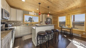 A kitchen with wooden ceiling, a central island with stools, and large windows offering a view.