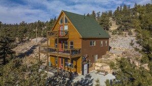 A two-story log cabin with a balcony, surrounded by trees and a rocky hillside.