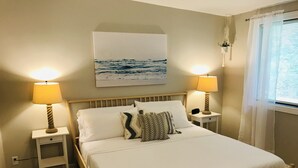 Ein ordentlich bezogenes Bett mit weißen Leinentüchern, zwei Nachttische mit Lampen, ein an der Wand hängendes Seebilder und ein Fenster mit durchscheinenden Vorhängen.