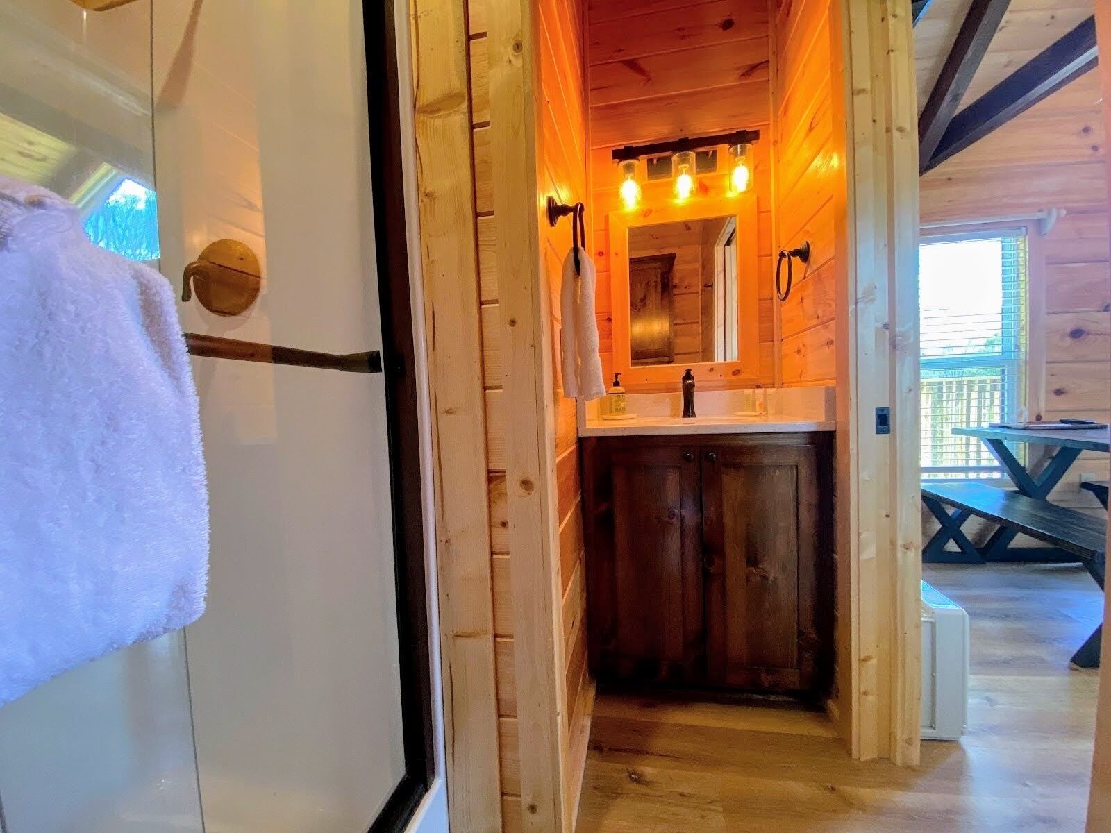 Interior de madera con un baño que incluye un lavabo, espejo e iluminación. Una toalla cuelga de una puerta, y se ve una mesa de picnic al fondo.