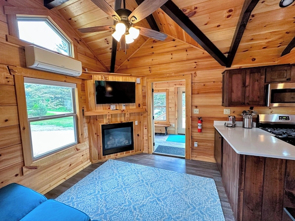 A cozy cabin interior with a fireplace, wooden walls, a ceiling fan, a TV, and a kitchen area.