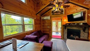 A wooden cabin interior with a sofa, table, fireplace, TV, and large windows. - B11 Awesome "Tiny Home" A/C, Mountain Views, Minutes to Skiing, Hiking, Attractions, Dog Friendly! (Twin Mountain)