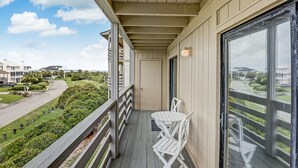 A balcony with a table and chairs, a door, and a view of a street and greenery. - Family friendly condo, Ocean views (Tybee Island)