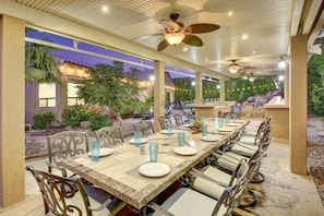 An outdoor dining area with a long table set for multiple people, surrounded by chairs with cushioned seats.