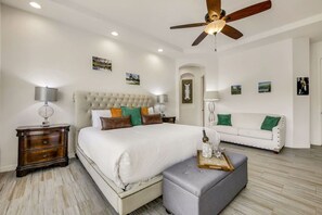 A bedroom with a large bed, a white sofa, a wooden nightstand, a ceiling fan, and wall-mounted artwork. - Echelon: Pool, Spa, Pool Table, Ping Pong table! (Indio)