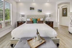 A bedroom with a large bed, two bedside tables, a nightstand, a window with blinds, and a wall-mounted mirror.