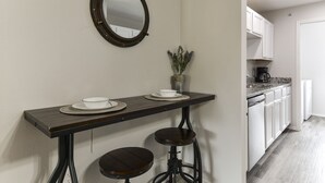 Una cocina con armarios blancos, una mesa de madera oscura con dos taburetes, un espejo redondo y un pequeño jarrón con flores secas.