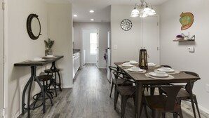 Un comedor con una mesa de madera dispuesta para seis personas, rodeada de taburetes de bar. Sobre la mesa cuelga una araña, y se observa un reloj de pared. La habitación tiene una paleta de colores neutros y suelo de madera clara.