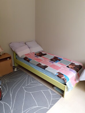 Quarto pequeno com uma cama de solteiro, moldura verde, colcha estampada e tapete floral cinza.