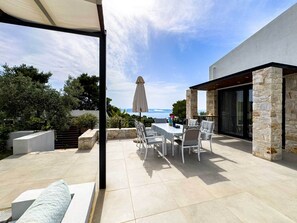 Een terras buiten met een eettafel en stoelen, een overdekte zithoek en uitzicht op de zee.