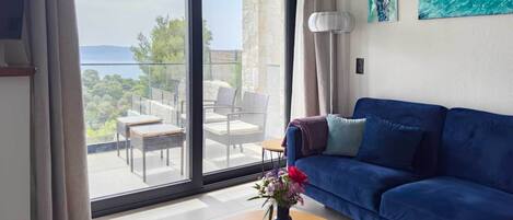 Un salon moderne avec un canapé bleu, une table basse ronde ornée d’un vase de fleurs et une vue sur un balcon aménagé avec du mobilier d’extérieur.