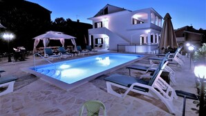 Una piscina con sillas de descanso y una zona de estar cubierta, rodeada de un patio de piedra y un edificio grande de color blanco con numerosas ventanas.