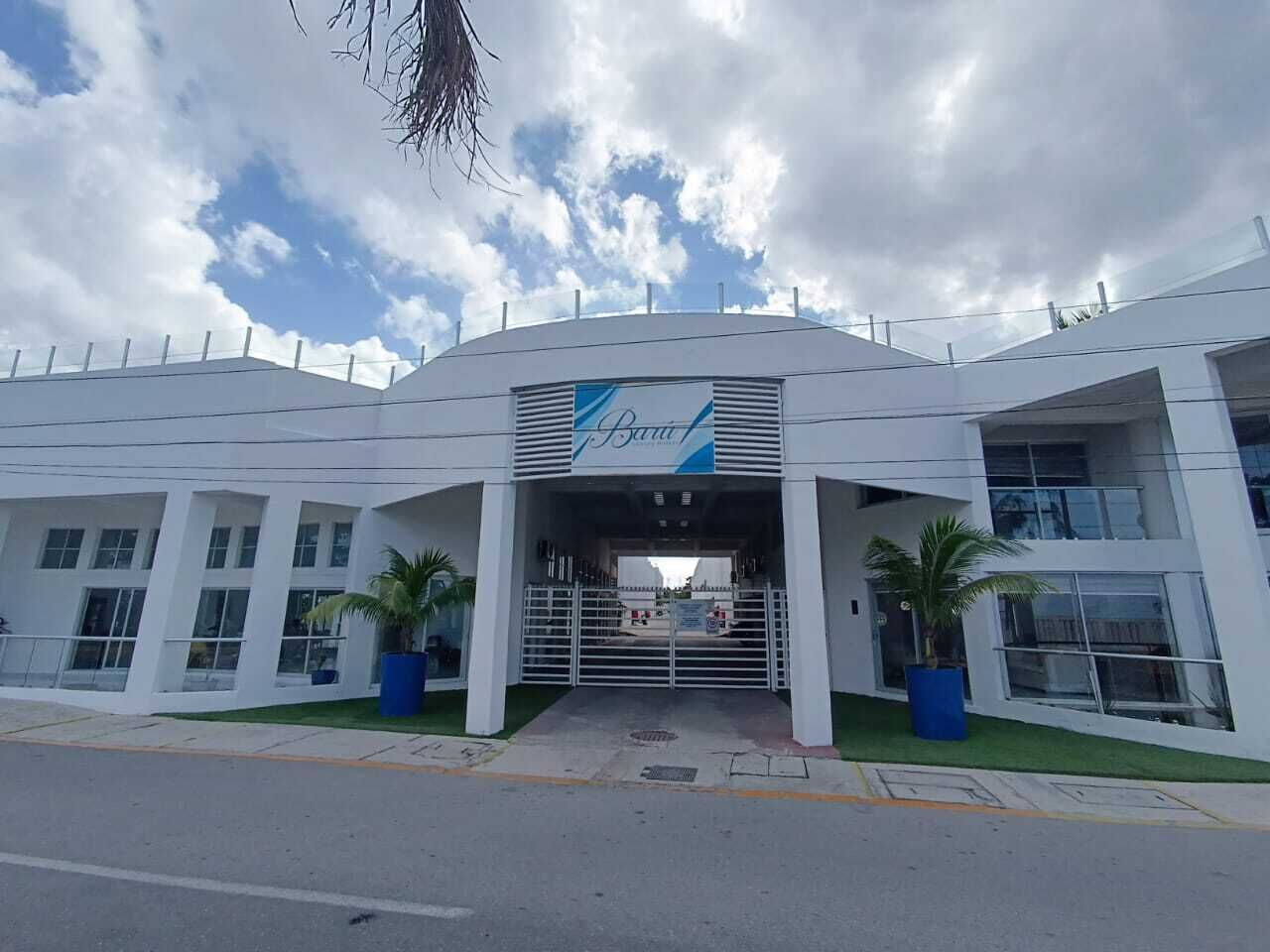 A modern building with a sign reading "Baril." It features a large entrance with a gated driveway, two potted palm trees, and a clear sky with clouds.
