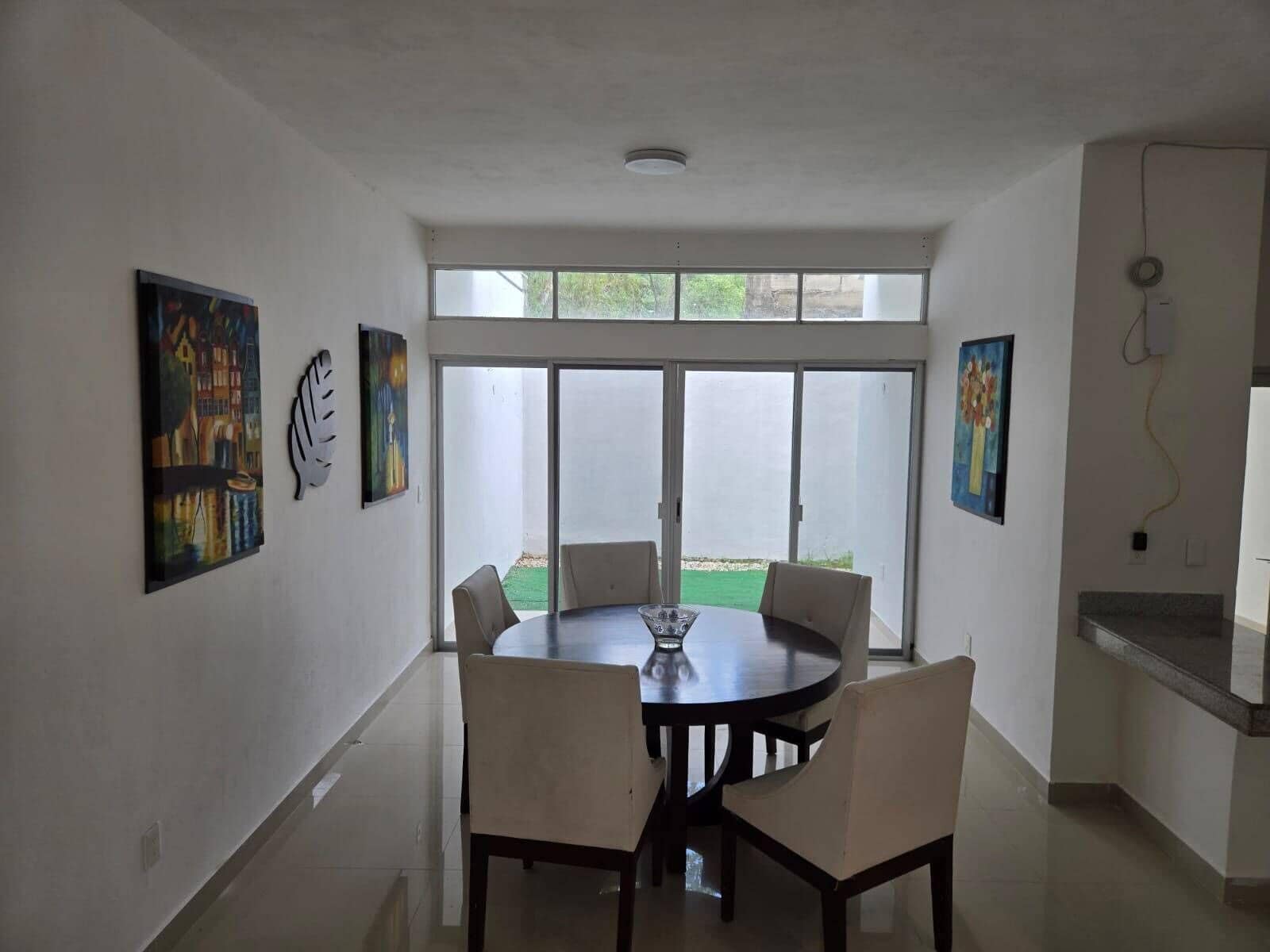 A dining area with a round table and chairs, large windows, and artwork on the walls.