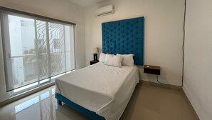 Une chambre moderne avec un grand lit, une tête de lit bleue, une fenêtre avec des stores et un climatiseur fixé au mur.