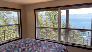 A bedroom with a bed and two large windows offering a view of a forest and the sea.