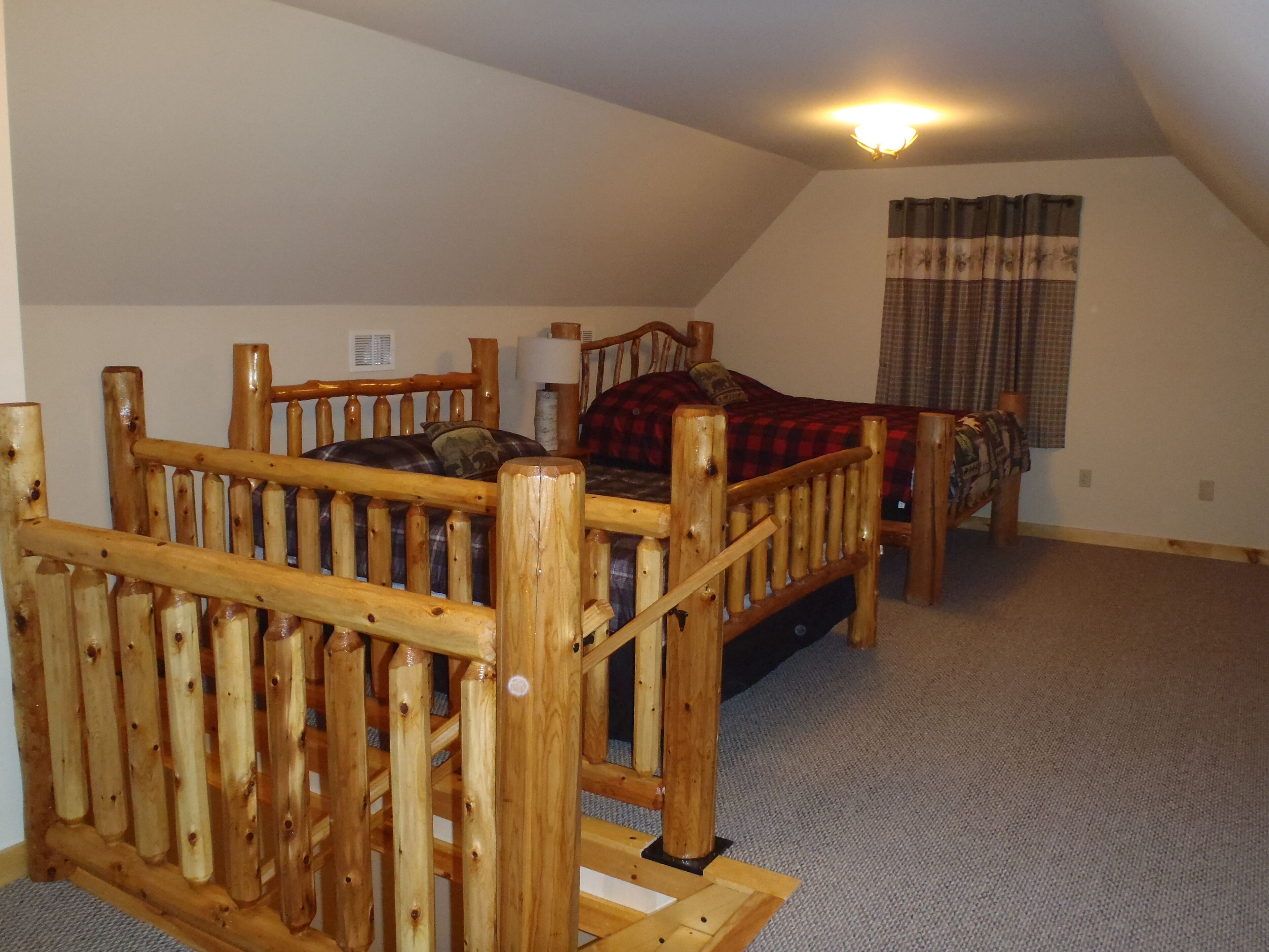 Ein Zimmer mit schrägen Wänden, einem hölzernen Bettgestell, einem Einzelbett mit kariertem Überwurf, einem Nachttisch und einer Wandlampe.