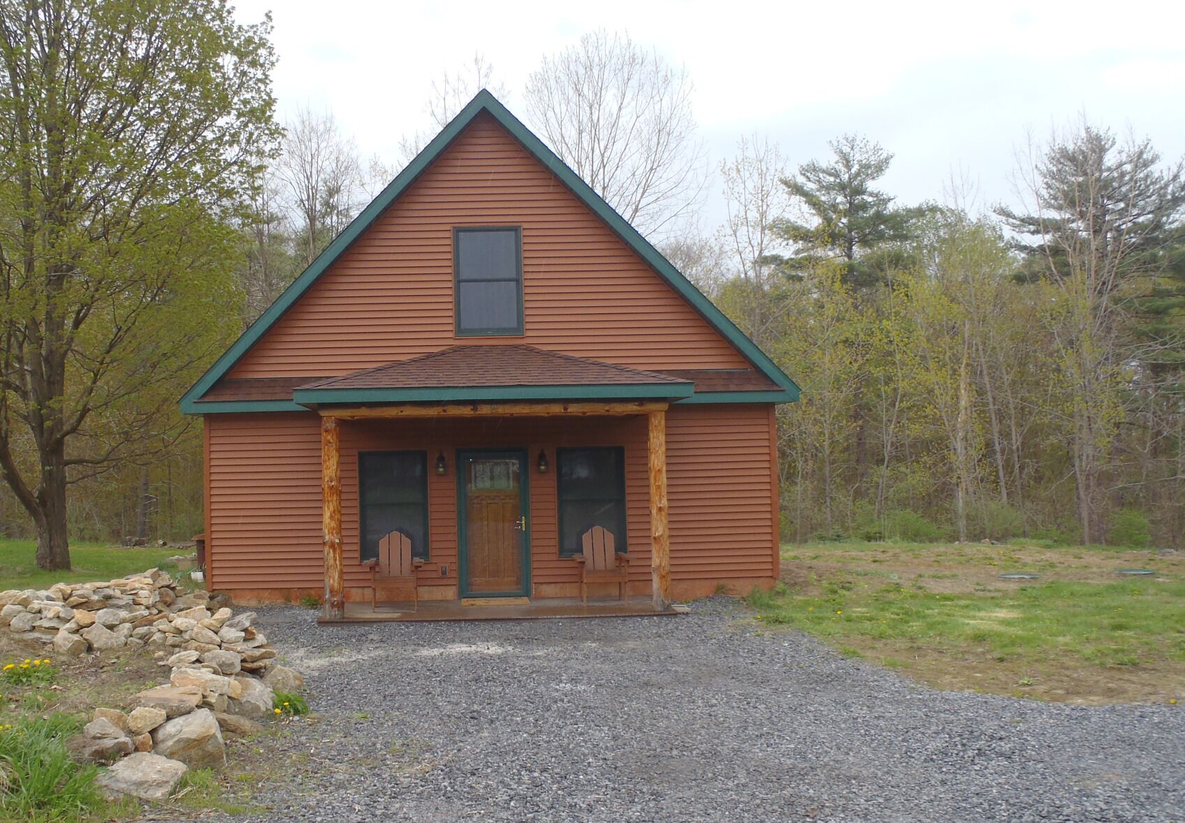 A wooden cabin with a gabled roof, a front porch with two chairs, and a gravel driveway.