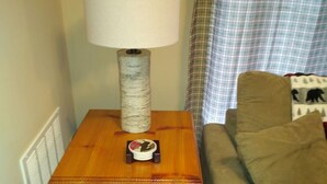 Une table en bois avec une lampe et un petit objet décoratif, un canapé marron et un rideau à carreaux dans une pièce équipée d’un radiateur et d’une fenêtre.