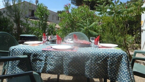 Un área de comedor al aire libre con una mesa dispuesta para cuatro personas, rodeada de follaje y un edificio de fondo.