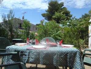 An outdoor dining area with a table set for four, surrounded by greenery and a building in the background. - Nice semi detached villa Mt St Martin Cap d'Agde (LE CAP D'AGDE)