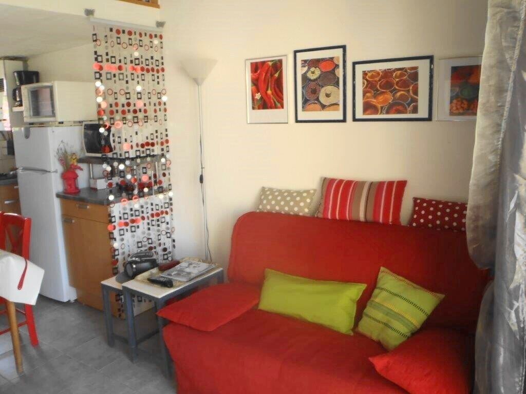 A compact living space with a red sofa, a small table, a kitchen area with a refrigerator, and a microwave. There are three framed artworks on the wall.