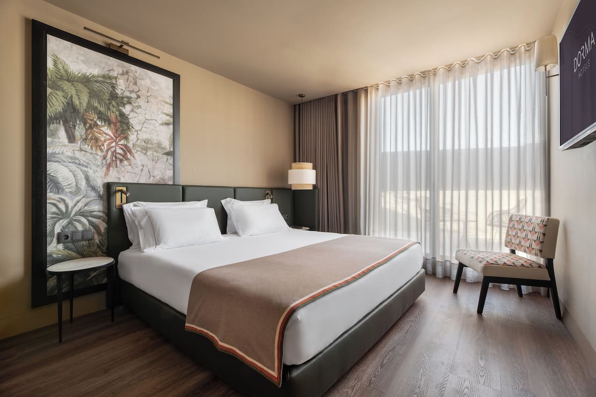 A hotel room with a bed, a chair, a nightstand, a lamp, and a painting on the wall.
