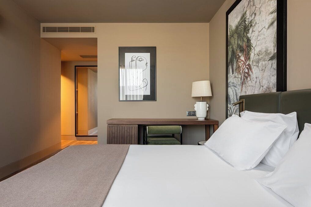 A hotel room with a bed, a desk with a lamp, a framed abstract artwork, and a door leading to another room.