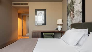 A hotel room with a bed, a desk with a lamp, a framed abstract artwork, and a door leading to another room.
