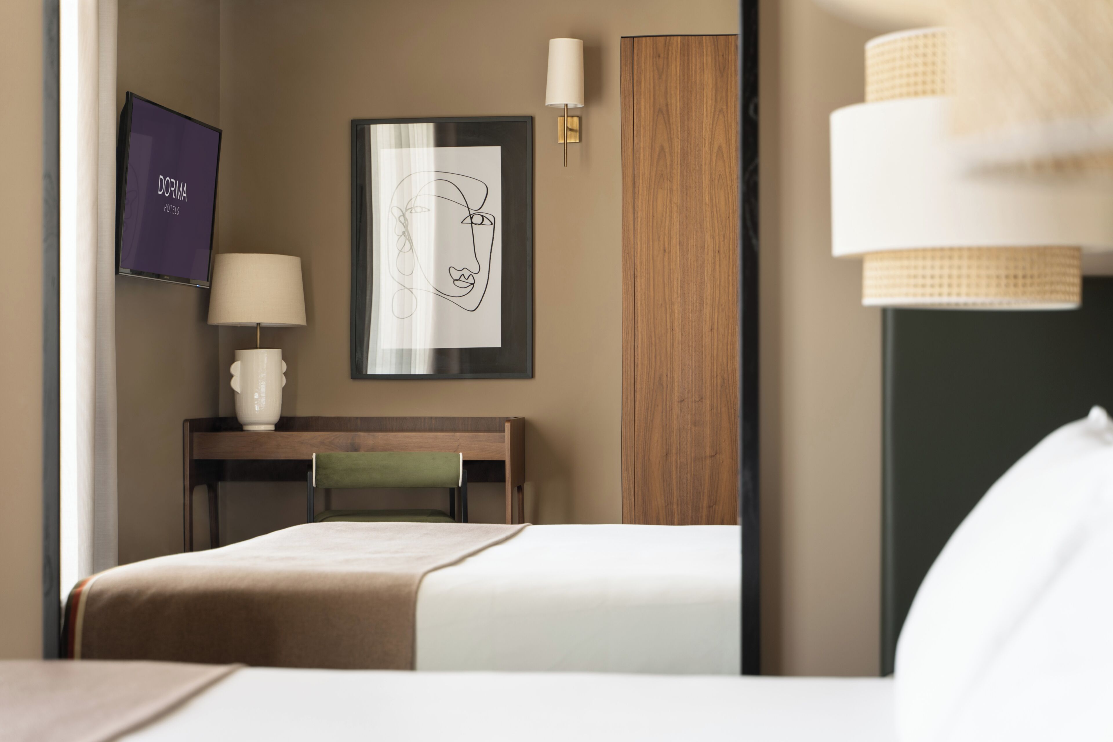 Et hotelværelse med en seng, et skrivebord, en stol, en lampe, et billede i ramme og et fjernsyn, der viser 'DORMA HOSTELS'.