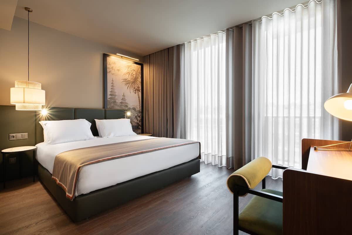 A modern hotel room with a large bed, a desk with a chair, a lamp, and a view of the outside through sheer curtains.