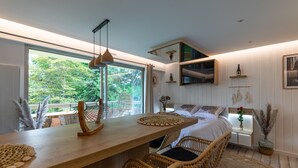 Een moderne kamer met een houten eettafel, rieten stoelen, een bed met wit beddengoed, een aan de muur bevestigde televisie en een groot raam met uitzicht op groen.
