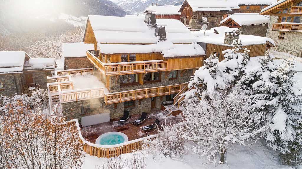 A multi-story wooden lodge with a hot tub and outdoor seating area, surrounded by snow-covered trees and mountains.