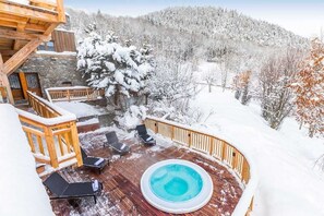 A wooden deck with a hot tub, lounge chairs, and a view of snow-covered trees and hills.